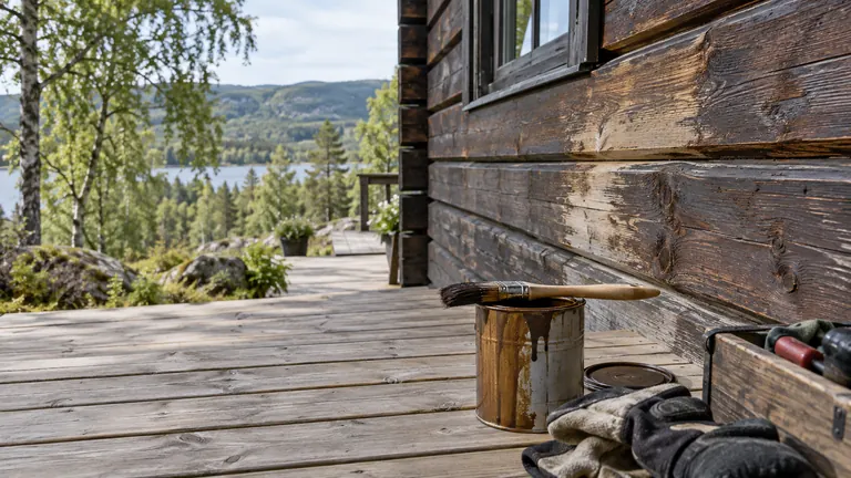 Beis, pensel og arbeidshansker ved en værslitt hyttevegg.
